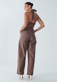 Bruine linnen jumpsuit met halternek en strikdetail, losse pasvorm, brede pijpen, zakken, gedragen met witte schoenen met hak. Achteraanzicht getoond.