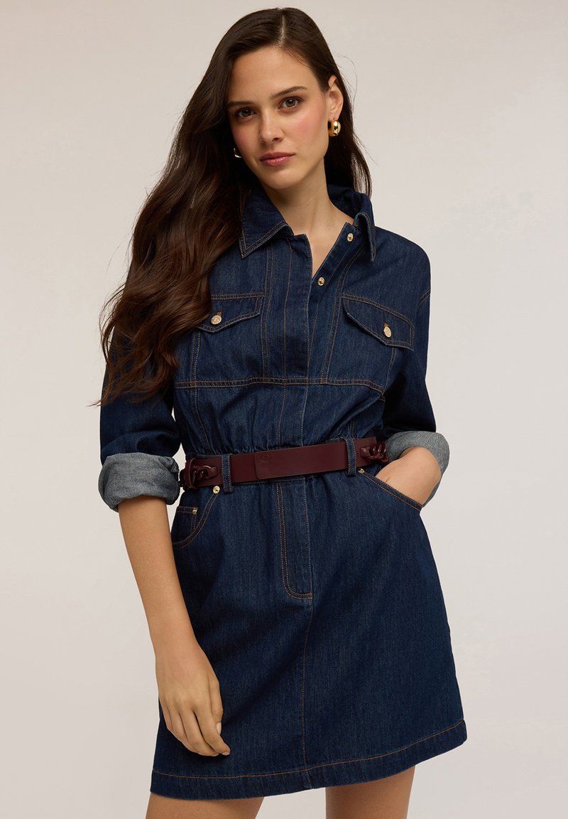 Robe chemise en denim avec une taille ceinturée, deux poches poitrine, de couleur bleu foncé, manches retroussées et détails de couture contrastants.