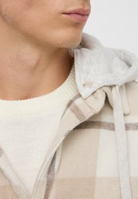 Giacca flanella a quadri beige con cappuccio grigio; presenta una chiusura a bottoni, una consistenza morbida e un maglione crema a coste sotto.