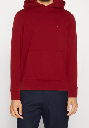 Mężczyzna w czerwonej bluzie z kapturem, z kieszenią z przodu i małym logo, w połączeniu z ciemnoniebieskimi spodniami, na jednolitym tle.