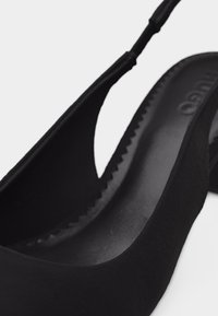 Μαύρο παπούτσι από σουέντ με λουράκι στο πίσω μέρος, διακοσμημένο με κυματιστή λεπτομέρεια και χαμηλό τετράγωνο τακούνι, που δείχνει την υφή της εσωτερικής σόλας και το λουράκι.