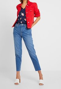 Giacca di jeans rossa, top floreale blu navy, jeans a vita alta di colore azzurro chiaro e scarpe con tacco aperte bianche con un design testurizzato.