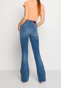 Vaqueros acampanados de mezclilla azul de talle alto con dos bolsillos traseros y un sutil desgaste, combinados con una camiseta naranja de manga corta.