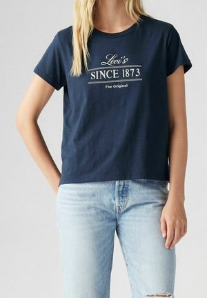 Person trägt ein marineblaues Levi's T-Shirt mit dem Schriftzug "SINCE 1873 The Original" und hellblaue Jeans, steht vor einem einfarbigen Hintergrund.