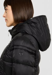 Zwarte gewatteerde jas met een hoge kraag, ritssluiting aan de voorkant en een afneembare capuchon. Heeft een doorgestikt patroon en een logodetail op de mouw.