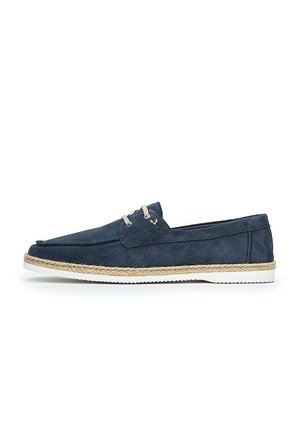 Zapato náutico de ante azul marino con puntera redondeada, suela de goma blanca y detalles de yute a lo largo del borde. Presenta dos ojales y costuras sutiles.