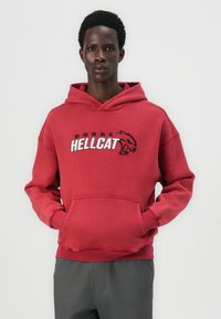 Roter Kapuzenpullover mit einer Fronttasche. Hat ein "DODGE HELLcat"-Grafik in Schwarz und Weiß. Hergestellt aus weichem, strukturiertem Stoff.