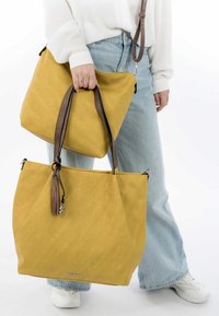 Geel canvas schoudertas met bruine leren handvatten, voorzien van een kwastaccent en een bijpassende kleinere tas. Gladde textuur, rechthoekige vorm.