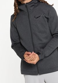 Veste zippée Nike gris foncé en tissu texturé, avec un col haut et un petit logo sur la poitrine gauche.