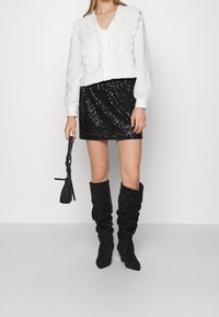 Vit blus med scallopkanter, svart paljetterad mini kjol och knähöga svarta stövlar med prydnader, håller en liten svart väska.