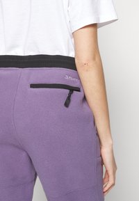 Lila fleece-joggers med svart elastiskt midjeband och en dragkedjeficka på baksidan. Har en liten logotyp på fickområdet.