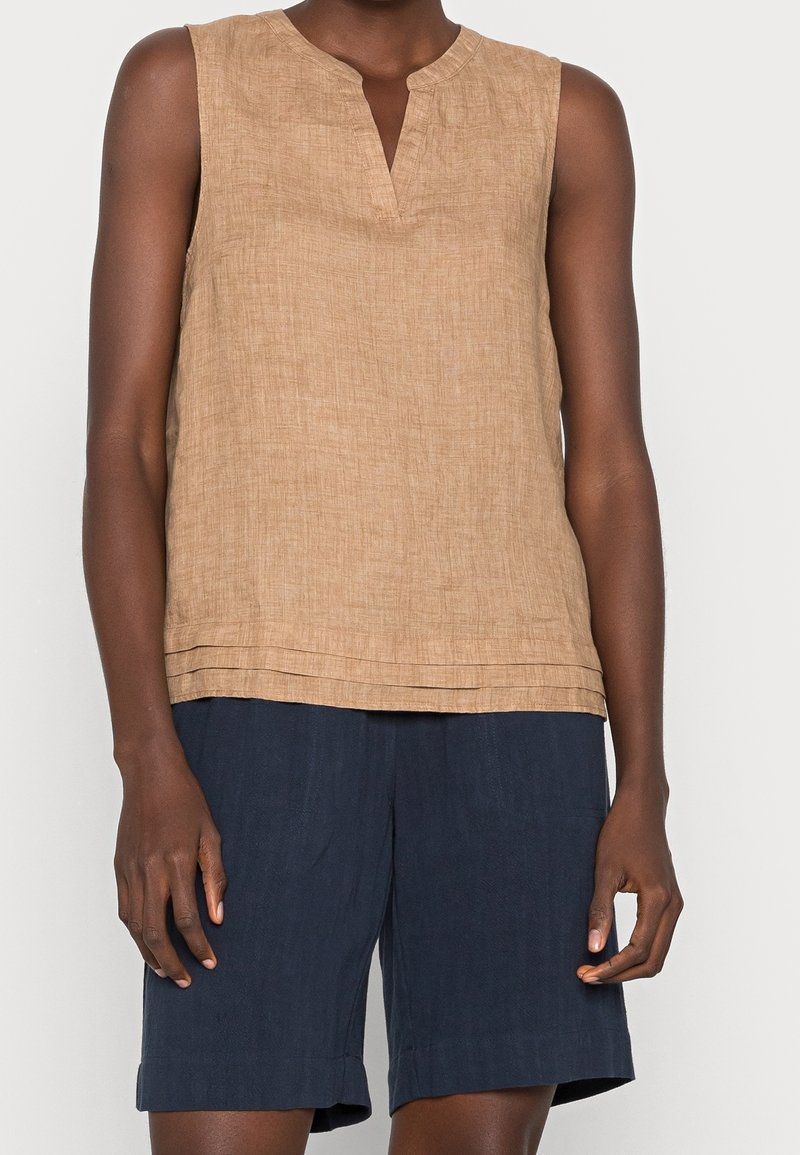 Haut sans manches en lin beige avec encolure crantée, associé à un short bleu marine longueur genou porté par une personne debout sur un fond uni.