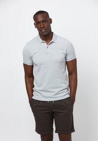 Szara koszulka polo z krótkim rękawem i kołnierzykiem zapinanym na dwa guziki, w połączeniu z ciemno-brązowymi szortami. Model stoi z rękami w kieszeniach, na neutralnym tle.