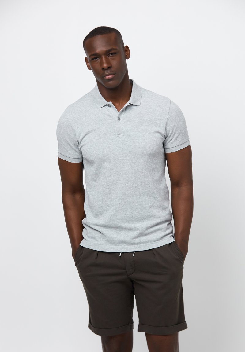 Szara koszulka polo z krótkim rękawem i kołnierzykiem zapinanym na dwa guziki, w połączeniu z ciemno-brązowymi szortami. Model stoi z rękami w kieszeniach, na neutralnym tle.