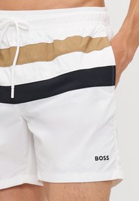 Costume de baie alb cu dungi orizontale în nuanțe de bej și bleumarin. Talie elastică cu șnur și mic logo negru "BOSS" pe lateral.