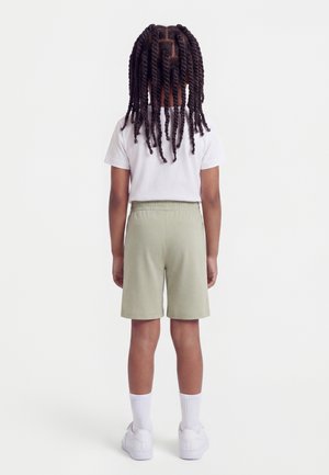 Copil cu păr lung răsucit, văzut de spate, purtând un tricou alb, pantaloni scurți bej, șosete albe și adidași albi, pe un fundal simplu.