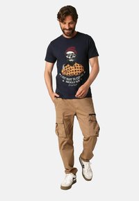 T-shirt graphique navy mettant en avant un homme barbu portant un bonnet rouge et une chemise à carreaux ; associé à un pantalon cargo beige et des baskets blanches.