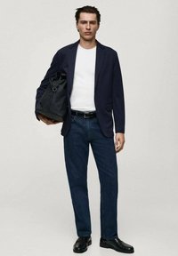 Marineblazer über einem weißen T-Shirt, dunkelblaue Jeans, schwarzer Gürtel und Schuhe. Hält eine schwarze Tasche mit gerolltem oberen Teil und steht vor einem hellen Hintergrund.