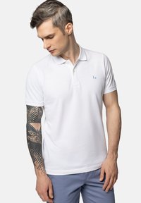 Biała bawełniana koszulka polo z klasycznym kołnierzykiem, krótkimi rękawami i małym niebieskim logo na lewej piersi. Gładka tekstura i dopasowany krój.