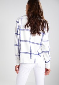 Sudadera blanca con diseño de cuadros azules, corte holgado, mangas largas y puños de canalé. Combinada con pantalones blancos para un look relajado.