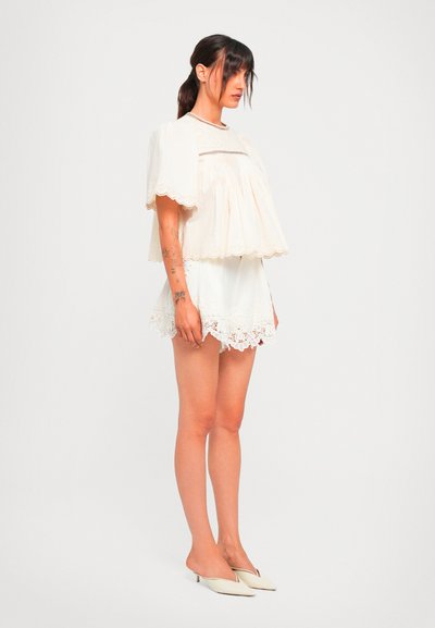 Woman with dark hair in a ponytail wearing a white lace-trimmed blouse and matching shorts, standing in beige pointed heels against a white background.