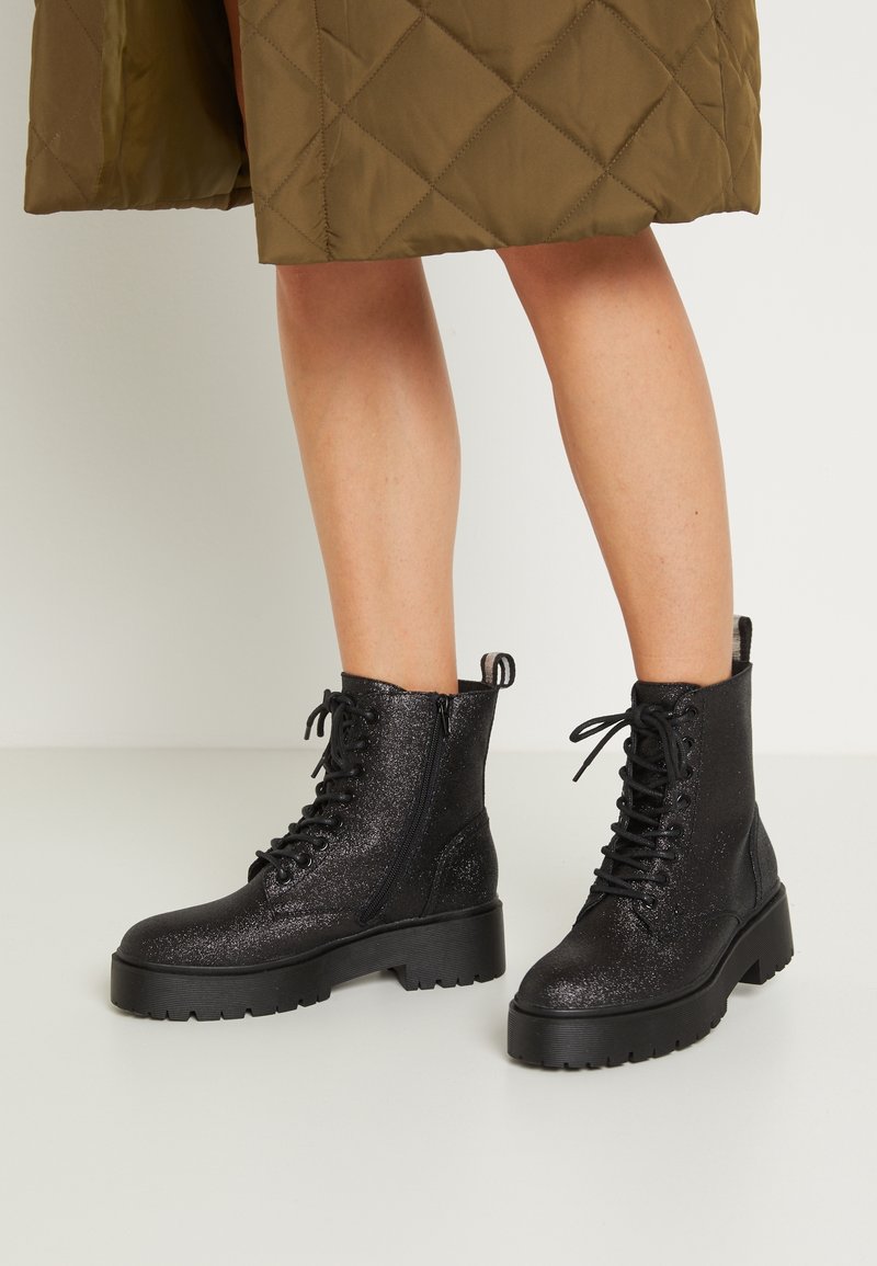 Bottines noires scintillantes à lacets avec fermetures éclair latérales portées avec une jupe matelassée vert olive, montrant les jambes et pieds sur un sol blanc.