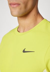 Lys gul atletisk T-shirt, kortærmet, rund halsudskæring, struktureret stof, med et sort Nike-logo på den øverste venstre side.