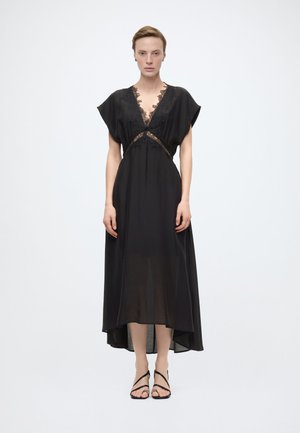 Femme portant une robe noire longueur cheville avec des détails en dentelle et des manches courtes, associée à des sandales noires à brides, debout devant un fond blanc.