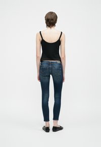 Persona de pie mirando hacia atrás, vestida con una blusa sin mangas negra, jeans ajustados oscuros y zapatos planos negros, sobre un fondo blanco liso.