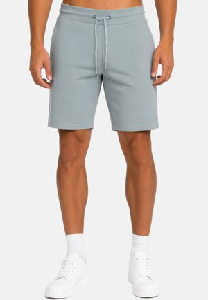 Uomo che indossa pantaloncini grigio chiaro con coulisse, calzini bianchi e sneakers bianche, in piedi davanti a uno sfondo uniforme.