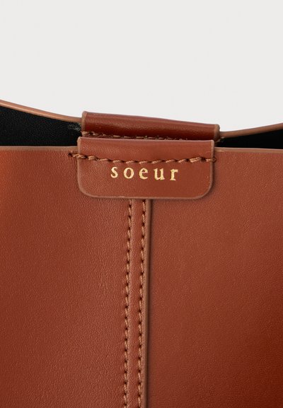 Hnedá kožená taška s elegantným dizajnom, textúrovaným povrchom, zlatým embosovaným logom "soeur" a viditeľným prešívaním pozdĺž šva.