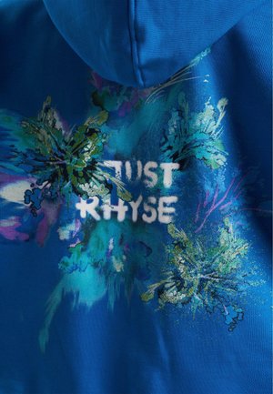 Blaues Hoodie mit einem lebhaften Blumenmuster in Grün und Lila, mit dem Text "JUST RHYSE" in Weiß über die Mitte.
