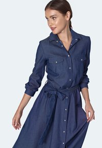 Sukienka koszulowa z denimu w ciemnoniebieskim kolorze, z zapięciem na guziki, dwiema kieszeniami na piersi, podwiniętymi rękawami i pasującym paskiem wiązanym w talii.