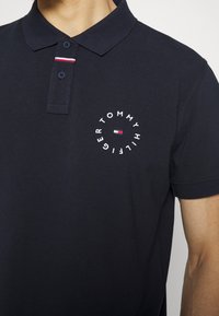 Mörkblå polo med två knappar, en liten röd, vit och blå rand nära kragen, och cirkulär Tommy Hilfiger-logotyp på bröstet.