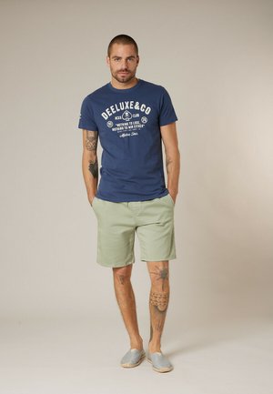 Homme aux cheveux courts et tatouages portant un t-shirt bleu à motif, un short vert clair et des chaussures grises sans lacets, debout les mains dans les poches.