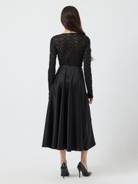 Kvinde iført en langærmet sort blondebluse og høj talje sort satin midi nederdel med sorte højhælede sko, set bagfra.