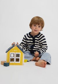 Kind, das barfuß auf dem Boden sitzt und mit einem gelben Spielhaus und Holzklötzen spielt, trägt einen gestreiften Pullover und Jeans.