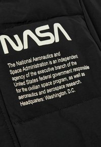 Tissu noir avec le logo "NASA" en blanc et des détails textuels décrivant la mission de l'organisation. Le texte est dans une police claire, sans empattement.