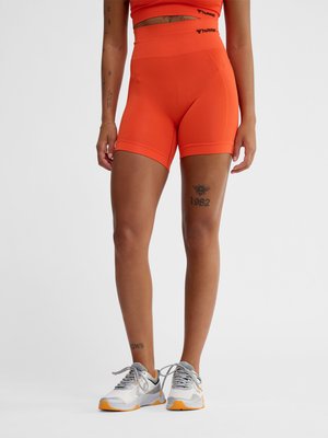 Persoon draagt feloranje biker shorts met hoge taille en een cropped top, met witte en oranje sportieve sneakers, zichtbare tatoeages op benen en armen.