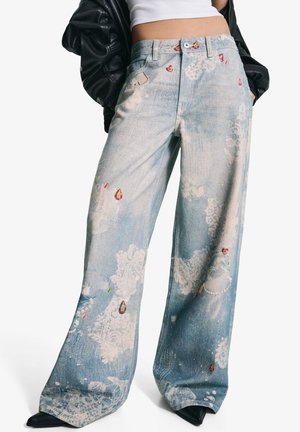 Jean large en denim avec un délavage bleu clair, des motifs floraux et des accents de type bijoux dans des teintes de rouge, vert et or.