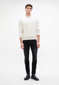 Suéter acanalado de color beige claro, vaqueros ajustados negros, botas de tobillo negras. El modelo está de pie en un fondo neutral, con los brazos relajados a los lados.