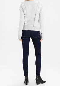 Kobieta stoi tyłem, ubrana w jasnoszary sweter z falbanami oraz ciemne dopasowane dżinsy i czarne botki na obcasie.