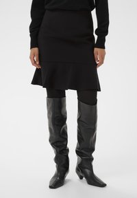 Black pencil skirt with a flared hemline, made of smooth fabric. Paired with form-fitting black leather knee-high boots featuring pointed toes.