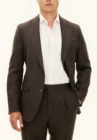 Giacca di lino marrone scuro con rever a punta, chiusura con un bottone e due tasche frontali, abbinata a una camicia chiara con colletto.