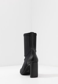 Bottine noire avec un talon bloc haut, possédant un dessus en tissu extensible et un dos en cuir lisse, caractérisée par des coutures minimales et un design épuré.