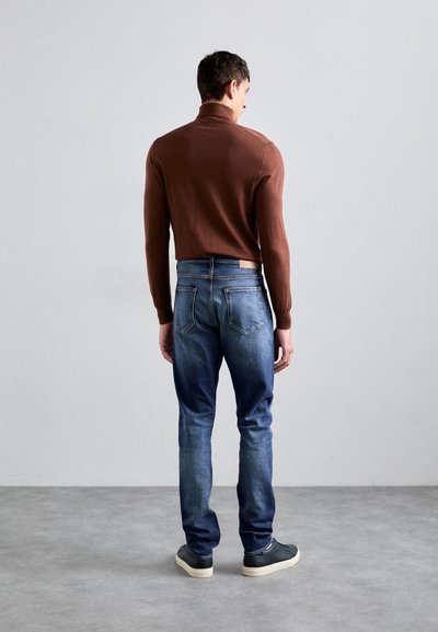 Pulôver castanho ajustado com mangas longas, combinado com jeans de ganga azuis. Os jeans têm um design clássico de cinco bolsos e um ligeiro desbotamento.