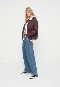 Giacca bomber color vino con colletto in shearling, abbinata a jeans a gamba larga blu chiaro e sneakers nere. Design semplice e moderno.