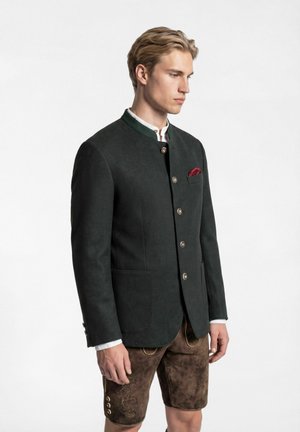 Junger Mann, der eine dunkle traditionelle Jacke mit silbernen Knöpfen und braune bestickte Lederhosen trägt, vor einem schlichten weißen Hintergrund stehend.