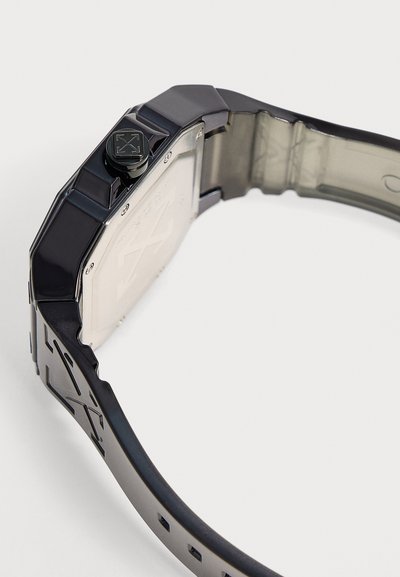 Montre-bracelet noire avec un bracelet à motif géométrique et un dos en métal, montrant une couronne latérale avec un motif de croix gravé.