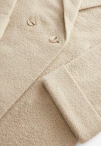 Manteau en mélange de laine beige clair avec une finition texturée, présentant un col cranté, deux boutons de couleur crème et des poignets repliés.